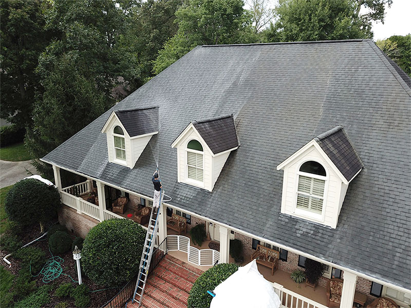technician applying soft wash roof treatment on residential asphalt shingle roof using low pressure system